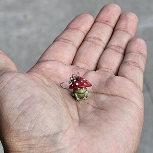 Puede incluir: Un pendiente detallado que representa una rana dorada con un gorro de seta rojo con lunares blancos. La rana est&aacute; sobre un aro plateado. El pendiente se sostiene en la palma de una mano abierta.