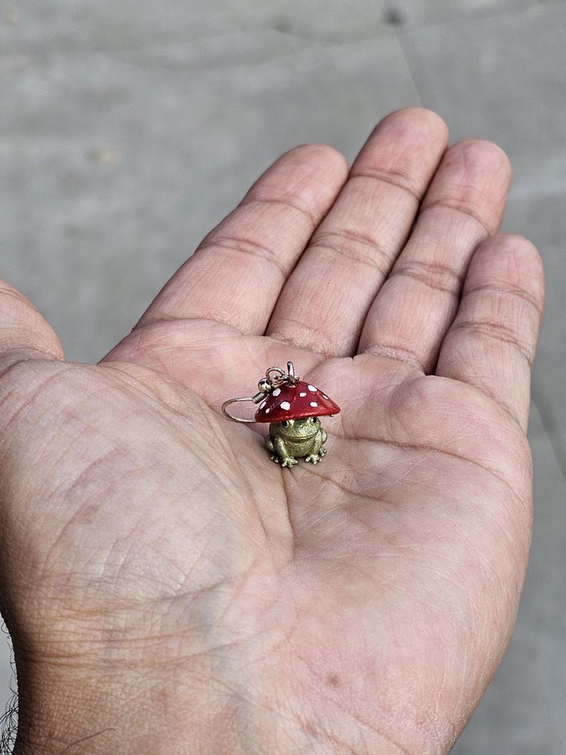 Puede incluir: Un pendiente detallado que representa una rana dorada con un gorro de seta rojo con lunares blancos. La rana est&aacute; sobre un aro plateado. El pendiente se sostiene en la palma de una mano abierta.
