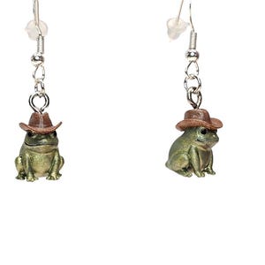 May include: A pair of novelty earrings featuring small, green frog figurines wearing brown cowboy hats. The frogs are suspended from silver-colored ear wires and chains, creating a whimsical accessory.