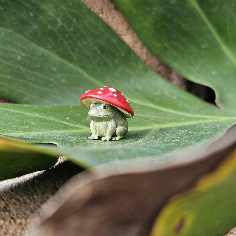 Puede incluir: Una peque&ntilde;a figura de rana verde con un gorro de seta roja con lunares blancos. La rana est&aacute; sentada en una hoja verde grande. Decoraci&oacute;n de jard&iacute;n caprichosa.