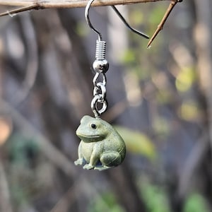 May include: A single silver earring with a frog charm. The frog is a muted green color with black eyes and sits on a chain. The earring is hanging from a thin, brown branch against a blurred background.