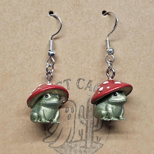 Puede incluir: Un par de pendientes caprichosos con peque&ntilde;as figuras de rana verde que llevan gorros de seta rojos con lunares blancos. Los pendientes cuelgan de ganchos plateados y se exhiben en una tarjeta marr&oacute;n.