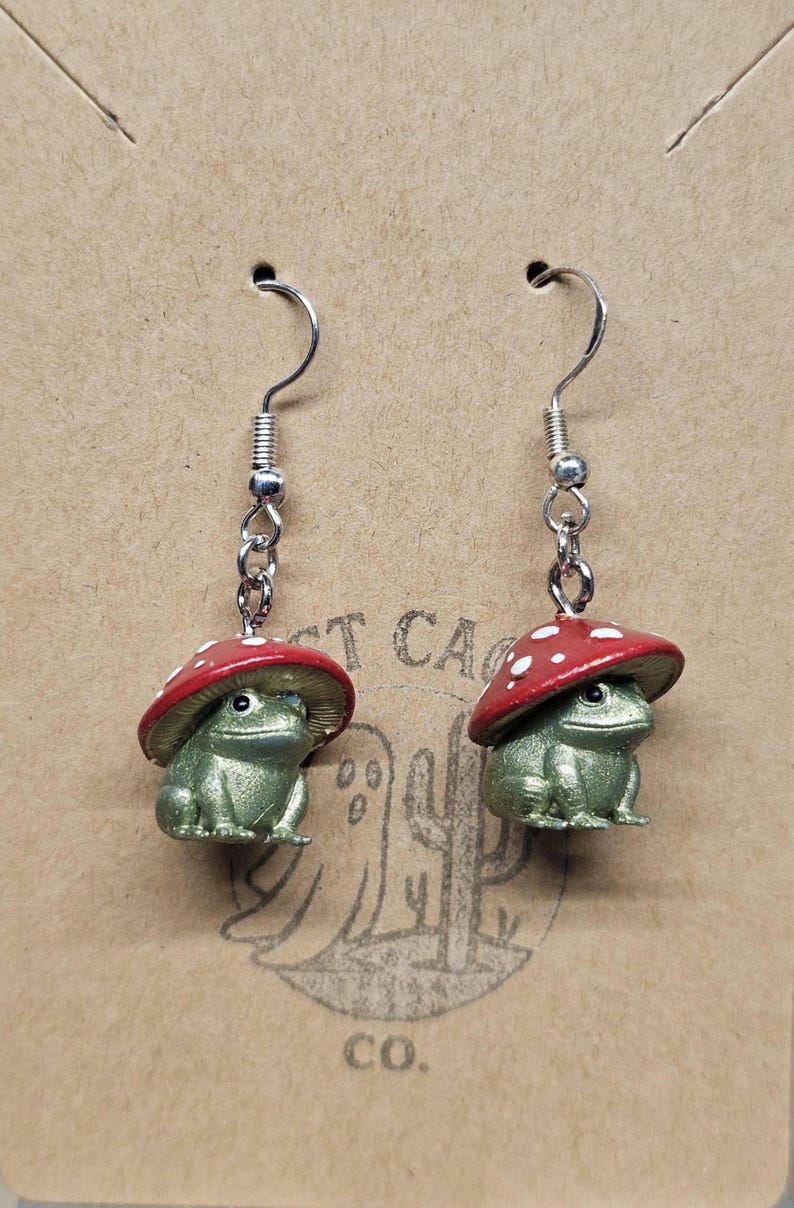 Puede incluir: Un par de pendientes caprichosos con peque&ntilde;as figuras de rana verde que llevan gorros de seta rojos con lunares blancos. Los pendientes cuelgan de ganchos plateados y se exhiben en una tarjeta marr&oacute;n.