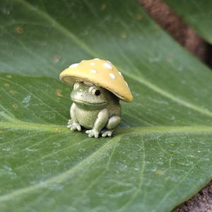 Puede incluir: Una peque&ntilde;a y detallada figura de una rana con un gorro de hongo amarillo con lunares blancos. La rana es de color verde apagado y est&aacute; sentada en una gran hoja verde. Una decoraci&oacute;n de jard&iacute;n caprichosa.