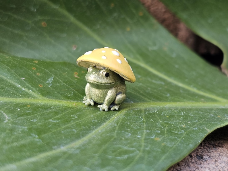 Puede incluir: Una peque&ntilde;a y detallada figura de una rana con un gorro de hongo amarillo con lunares blancos. La rana es de color verde apagado y est&aacute; sentada en una gran hoja verde. Una decoraci&oacute;n de jard&iacute;n caprichosa.
