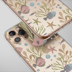 Może przedstawiać: Dwa przezroczyste etui na telefon z wzorem życia morskiego. Wzór przedstawia różowe i niebieskie ryby, rozgwiazdy i wodorosty na beżowym tle. Etui mają złote obramowanie.