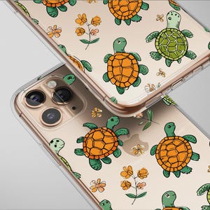 May include: Clear phone cases with a repeating pattern of cartoon turtles in orange and green, along with small yellow flowers and green leaves. The cases are shown at an angle, with the camera lenses visible.