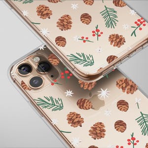 May include: Two clear phone cases featuring a festive design. The pattern includes brown pine cones, green pine needles, red berries, and white star accents on a beige background. The cases have a transparent border and a camera cutout.