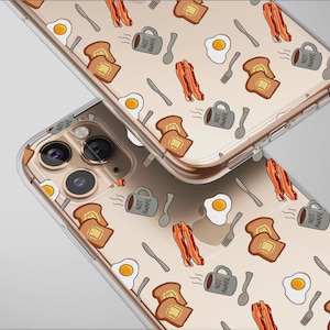 Könnte beinhalten: Zwei transparente Handyhüllen mit einem Frühstücksmuster. Das Design umfasst Illustrationen von Spiegeleiern, Speckstreifen, Toast, Kaffeetassen mit dem Text "NOT AWAKE", Löffeln und Gabeln. Der Hintergrund ist hellgrau.