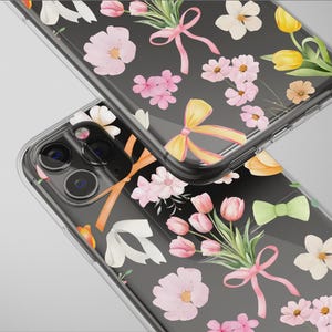 Puede incluir: Fundas transparentes para teléfono con diseño floral. Las fundas presentan flores rosas, blancas y amarillas, hojas verdes y lazos rosas y amarillos sobre un fondo negro. Las fundas están en ángulo, mostrando la cámara del teléfono.