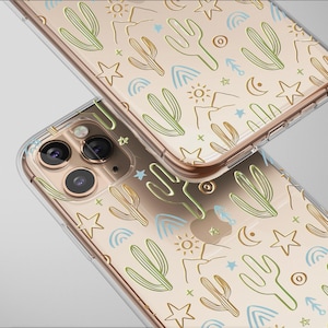 May include: Two clear phone cases with a desert-themed pattern. The design features cacti, stars, suns, moons, and mountains in green, brown, and blue on a beige background. The cases are made of clear plastic and have a gold-colored frame.