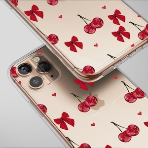 May include: Clear phone cases with a repeating pattern of red cherries, bows, and hearts. The cases are shown at an angle, displaying the camera lenses and the design on the back. The background is a light gray.