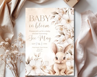 May include: A baby shower invitation with a watercolor design. The invitation features the text "BABY in bloom" and "Joe May". A cute bunny illustration is surrounded by white flowers. The invitation is on a white background with beige accents.