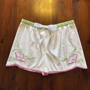 May include: White shorts with a green gingham waistband and a light blue ribbon tie. The shorts feature embroidered pink and blue flowers with green leaves and a pink lace trim along the bottom edge.