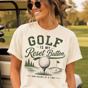 Puede incluir: Una camiseta de manga corta color crema con un estampado gráfico en verde oscuro. El texto dice GOLF IS MY Reset Button y ONE ROUND AT A TIME. El diseño incluye una pelota de golf sobre un tee, una bandera en el green y pinos.