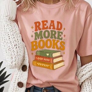 May include: A light pink t-shirt with the text "READ MORE BOOKS" in orange and green, with a stack of illustrated books. The shirt is worn with a white cardigan and blue jeans.