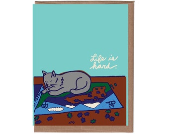 May include: A greeting card with a blue background and a gray cat sleeping on a puzzle. The text "Life is hard." is written in white on the card.