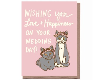 Può includere: Un biglietto di nozze rosa con il testo "Wishing you Love + Happiness on your Wedding Day!". Presenta due gatti dei cartoni animati con corone di fiori. I gatti sono grigi e bianchi e il testo è scritto a mano.