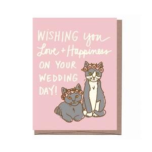 May include: A pink wedding card with the text "Wishing you Love + Happiness on your Wedding Day!" features two cartoon cats wearing flower crowns. The cats are gray and white, and the text is in a handwritten style.