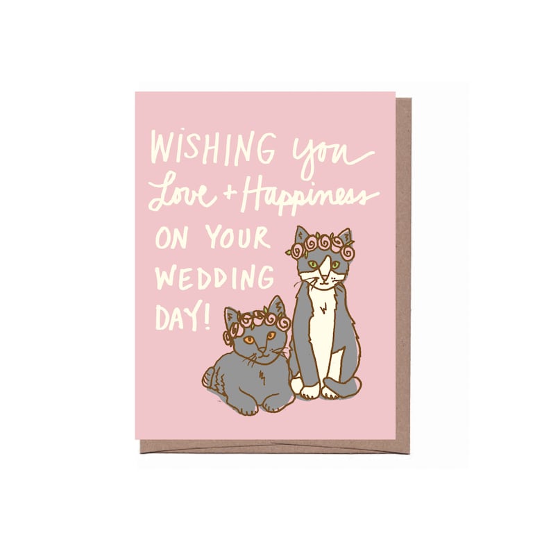 May include: A pink wedding card with the text "Wishing you Love + Happiness on your Wedding Day!" features two cartoon cats wearing flower crowns. The cats are gray and white, and the text is in a handwritten style.