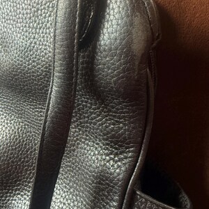 May include: A dark brown handbag featuring a textured, pebbled surface with visible stitching along the seams. The bag includes a vertical strap detail and a metallic silver-toned hardware ring attachment at the top corner of the main body.