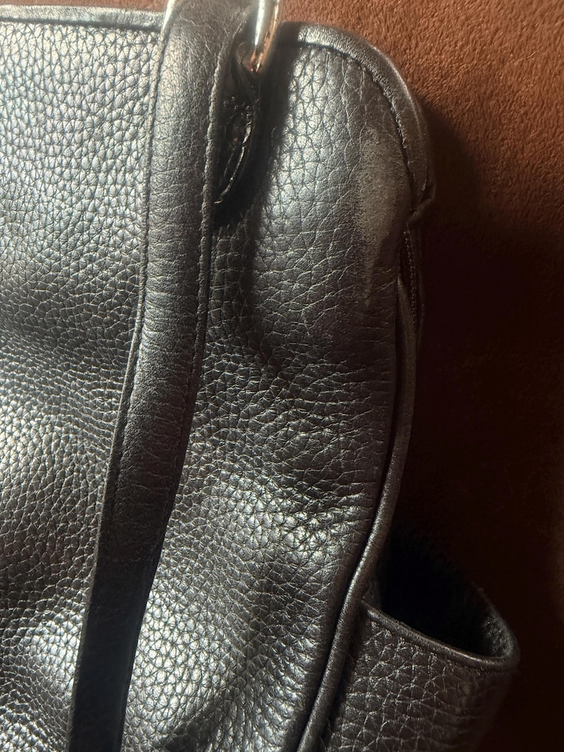 May include: A dark brown handbag featuring a textured, pebbled surface with visible stitching along the seams. The bag includes a vertical strap detail and a metallic silver-toned hardware ring attachment at the top corner of the main body.