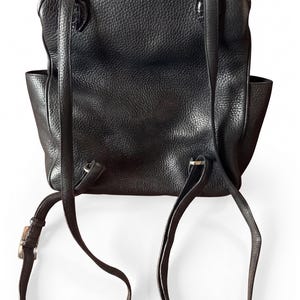 May include: A black backpack with a textured surface, featuring two adjustable shoulder straps attached with silver-toned metal hardware. The bag has a rectangular main compartment with side pockets and a flap closure secured by metal rings.