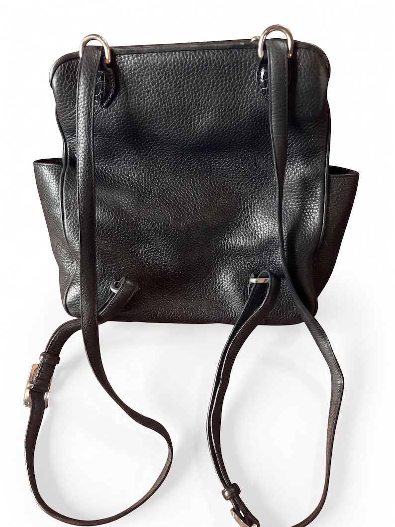 May include: A black backpack with a textured surface, featuring two adjustable shoulder straps attached with silver-toned metal hardware. The bag has a rectangular main compartment with side pockets and a flap closure secured by metal rings.