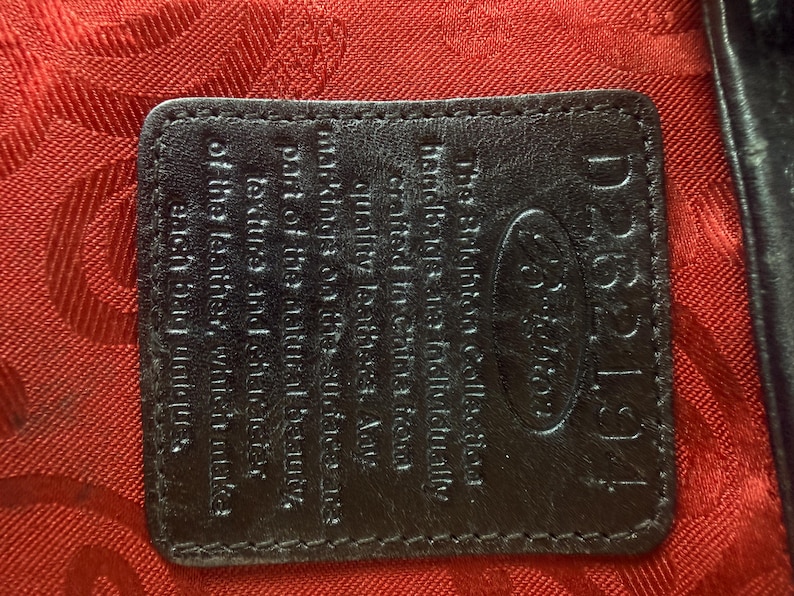May include: A dark brown leather rectangular patch with rounded corners, embossed with the Brighton logo and text, stitched onto a vibrant red fabric lining featuring a repeating tonal pattern. The text reads: The Brighton Collection. Handbags are individually crafted in China from quality leather. Any markings on the surface are part of the natural beauty, texture and character of the leather, which make each bag unique.