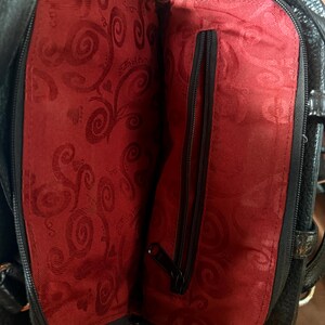 May include: A black handbag with a textured exterior and a vibrant red interior lining. The interior features a repeating swirl pattern and a central vertical zippered pocket with a black zipper pull, providing a contrast to the black outer shell.