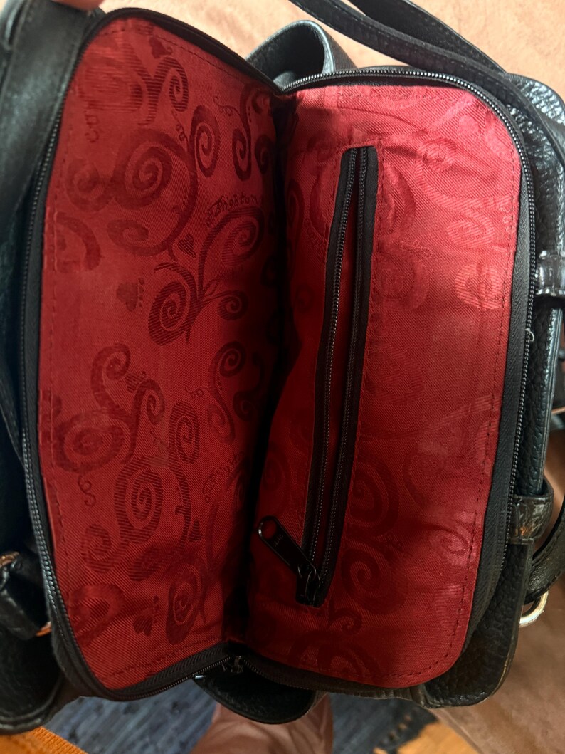 May include: A black handbag with a textured exterior and a vibrant red interior lining. The interior features a repeating swirl pattern and a central vertical zippered pocket with a black zipper pull, providing a contrast to the black outer shell.