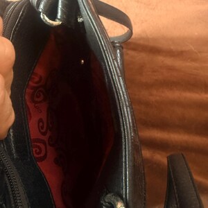 May include: A black handbag with a textured exterior and a vibrant red interior lining featuring a repeating black swirl pattern. The bag includes a zippered closure, silver-toned metal hardware, and a black strap attached with metal rings.