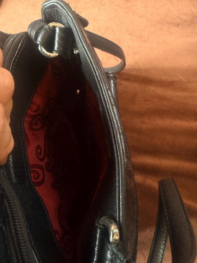 May include: A black handbag with a textured exterior and a vibrant red interior lining featuring a repeating black swirl pattern. The bag includes a zippered closure, silver-toned metal hardware, and a black strap attached with metal rings.
