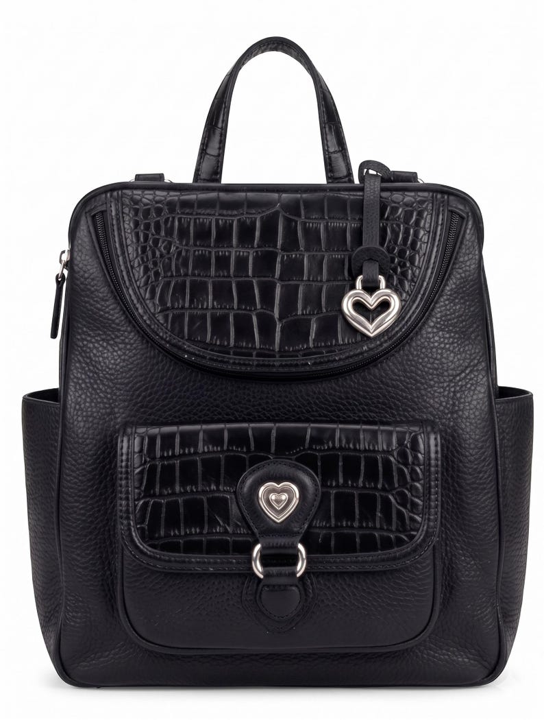 May include: A black backpack featuring a combination of pebbled and crocodile-embossed textures. The bag includes a top handle, a front pocket with a silver heart-shaped emblem, and a matching silver heart charm hanging from the upper zipper.