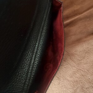 May include: A black textured handbag with a flap closure. The interior lining is a deep red color and features a repeating embossed pattern throughout the fabric. The exterior surface of the bag has a pebbled grain texture and a stitched seam.