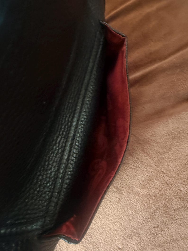 May include: A black textured handbag with a flap closure. The interior lining is a deep red color and features a repeating embossed pattern throughout the fabric. The exterior surface of the bag has a pebbled grain texture and a stitched seam.