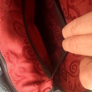 May include: The interior of a dark bag featuring a vibrant red lining with a repeating dark red swirl pattern. A black zippered pocket is integrated into the side of the bag, providing a secure compartment within the main storage area.