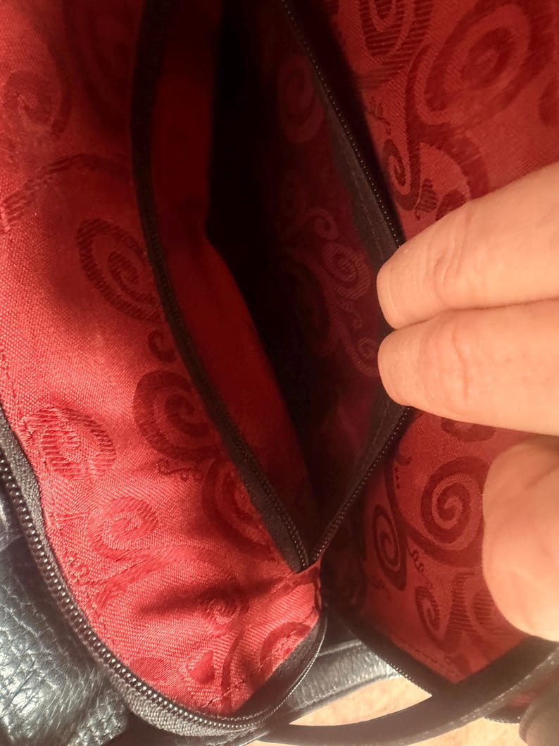 May include: The interior of a dark bag featuring a vibrant red lining with a repeating dark red swirl pattern. A black zippered pocket is integrated into the side of the bag, providing a secure compartment within the main storage area.