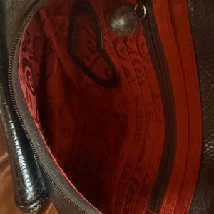 May include: A black leather bag with a textured exterior and a vibrant red interior lining. The interior features a zippered pocket, multiple vertical card slots, and a heart-shaped cutout detail on the fabric. A small heart-shaped metal charm is attached to the zipper pull.