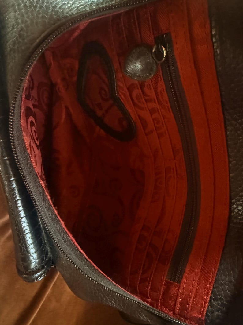May include: A black leather bag with a textured exterior and a vibrant red interior lining. The interior features a zippered pocket, multiple vertical card slots, and a heart-shaped cutout detail on the fabric. A small heart-shaped metal charm is attached to the zipper pull.