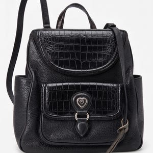 May include: A black backpack featuring a pebbled leather texture with crocodile-embossed leather accents on the top flap and front pocket. The front pocket includes a silver-toned heart-shaped emblem and a dangling heart-shaped metal charm.
