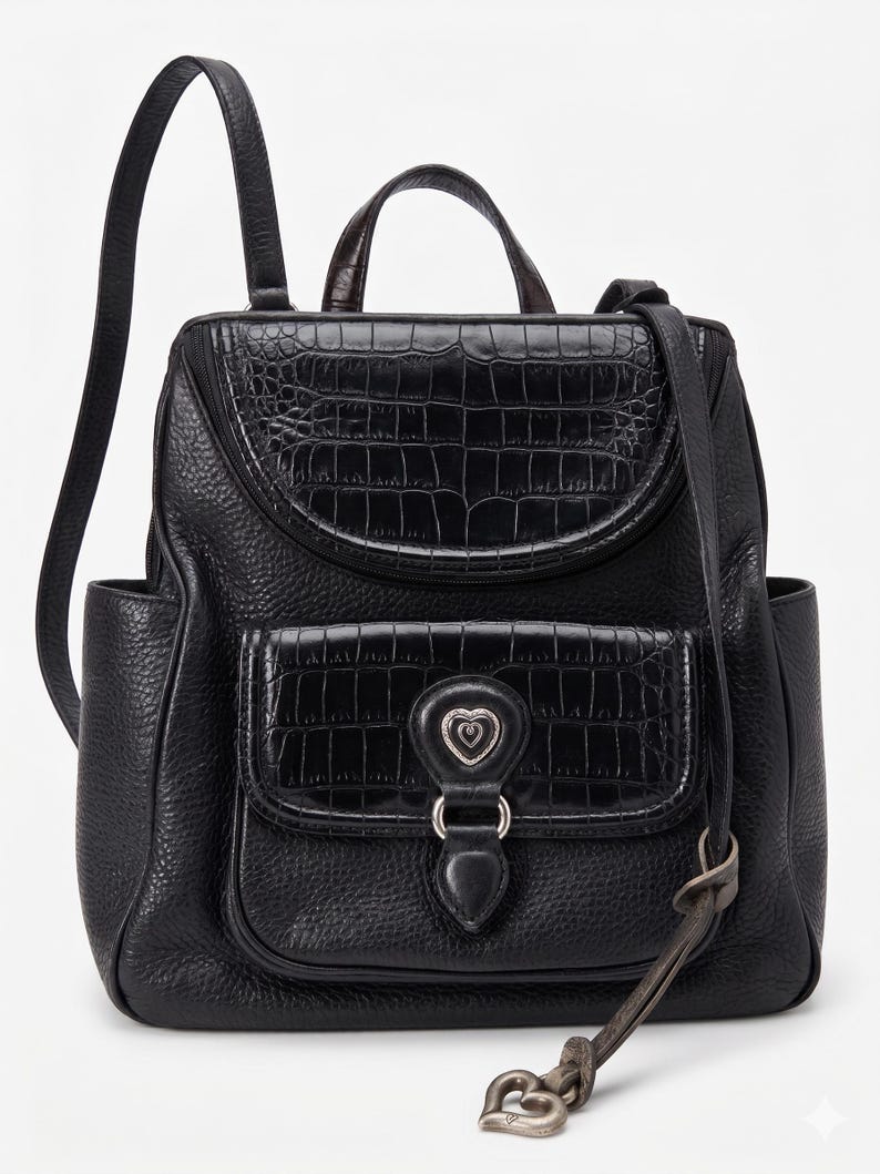 May include: A black backpack featuring a pebbled leather texture with crocodile-embossed leather accents on the top flap and front pocket. The front pocket includes a silver-toned heart-shaped emblem and a dangling heart-shaped metal charm.