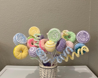 May include: A colorful bouquet of handmade, fabric-covered lollipops, donuts, and spiral decorations in a patterned ceramic vase. The lollipops and donuts are in pastel colors like yellow, pink, blue, and purple. The vase has a black and white chevron design.