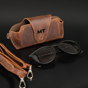 May include: A brown leather glasses case with the initials "MT" embossed on the front, next to a pair of black sunglasses. A matching leather strap with silver clasps is also visible. The case and glasses are on a black surface.