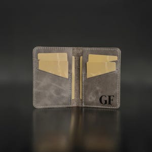 Könnte beinhalten: Offenes graues Lederportemonnaie mit mehreren Kartenfächern. Das Portemonnaie ist geöffnet und zeigt mehrere goldfarbene Karten. Die Initialen "GF" sind in Schwarz unten rechts aufgedruckt.