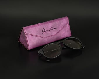 May include: A pair of black sunglasses next to a textured, triangular, magenta glasses case. The glasses case has the name "Flower Harper" in cursive script. The sunglasses have dark lenses and a black frame. The background is black.