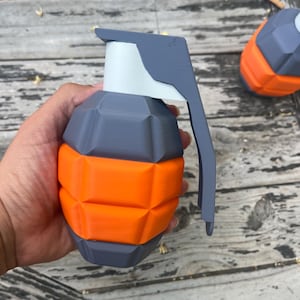 May include: A hand grenade replica featuring a geometric, faceted design with alternating horizontal bands of dark gray and bright orange plastic. The top assembly is a solid dark gray lever and a white cylindrical neck, creating a distinct contrast.
