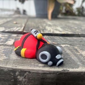 May include: A small figurine of a character wearing a red suit with black accents, a black mask with circular gray eye lenses, and a yellow-tipped gray tank strapped to the back. The figure is in a prone position with yellow bands on the arms.