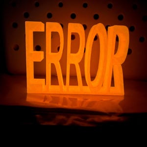 Könnte beinhalten: Ein freistehendes, dreidimensionales Schild mit dem Wort ERROR in fetten Großbuchstaben. Das Objekt strahlt ein helles, warmes orangefarbenes Licht aus, das einen sanften Schein auf die darunter liegende Fläche wirft, mit einer dunklen Lochplatte dahinter.