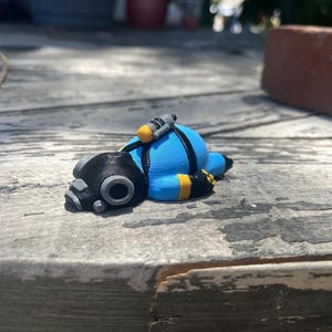 Puede incluir: Una pequeña figura de un buceador con traje azul brillante, casco negro, arnés negro y detalles amarillos. El buceador está tumbado en posición prona, con una pequeña botella de oxígeno amarilla en la espalda y una máscara negra detallada.